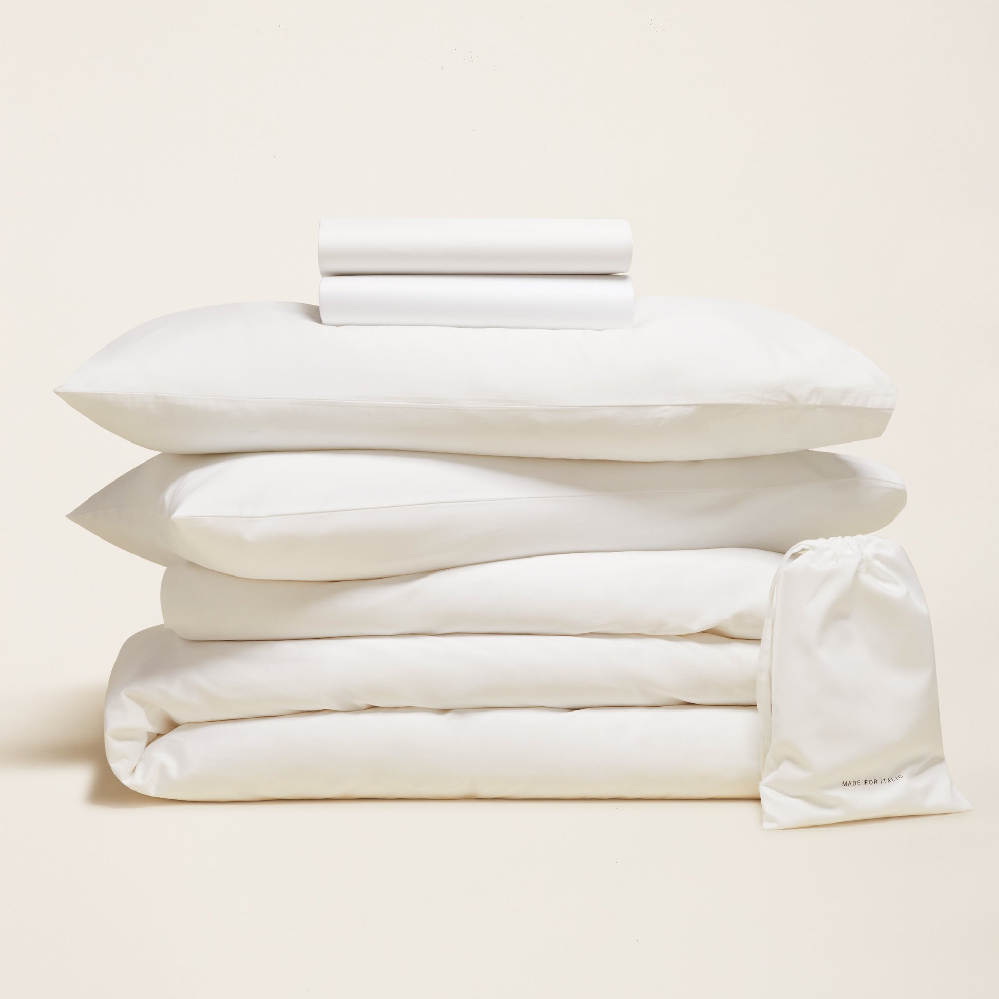 Stack of white bedding including a duvet, pillows, and a comforter on a beige background.