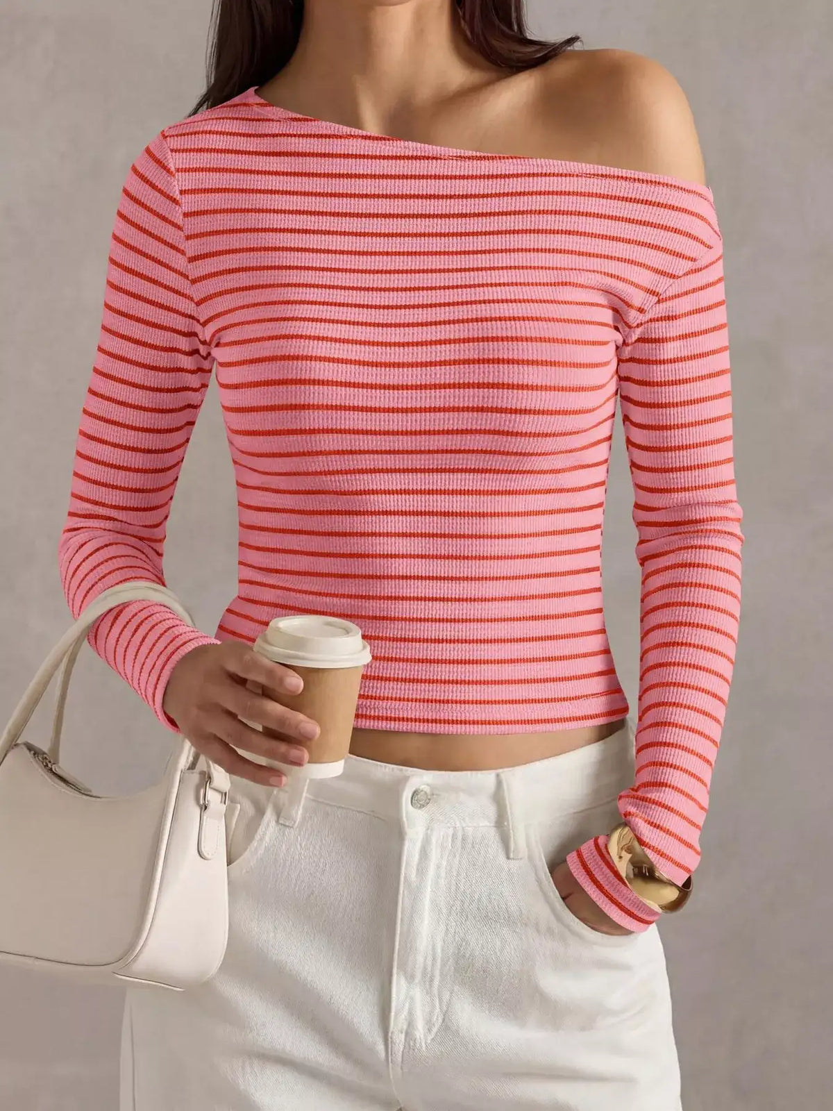 A woman wearing a coral pink and white horizontally striped off-the-shoulder long-sleeve crop top, paired with white pants and holding a white handbag and a coffee cup.