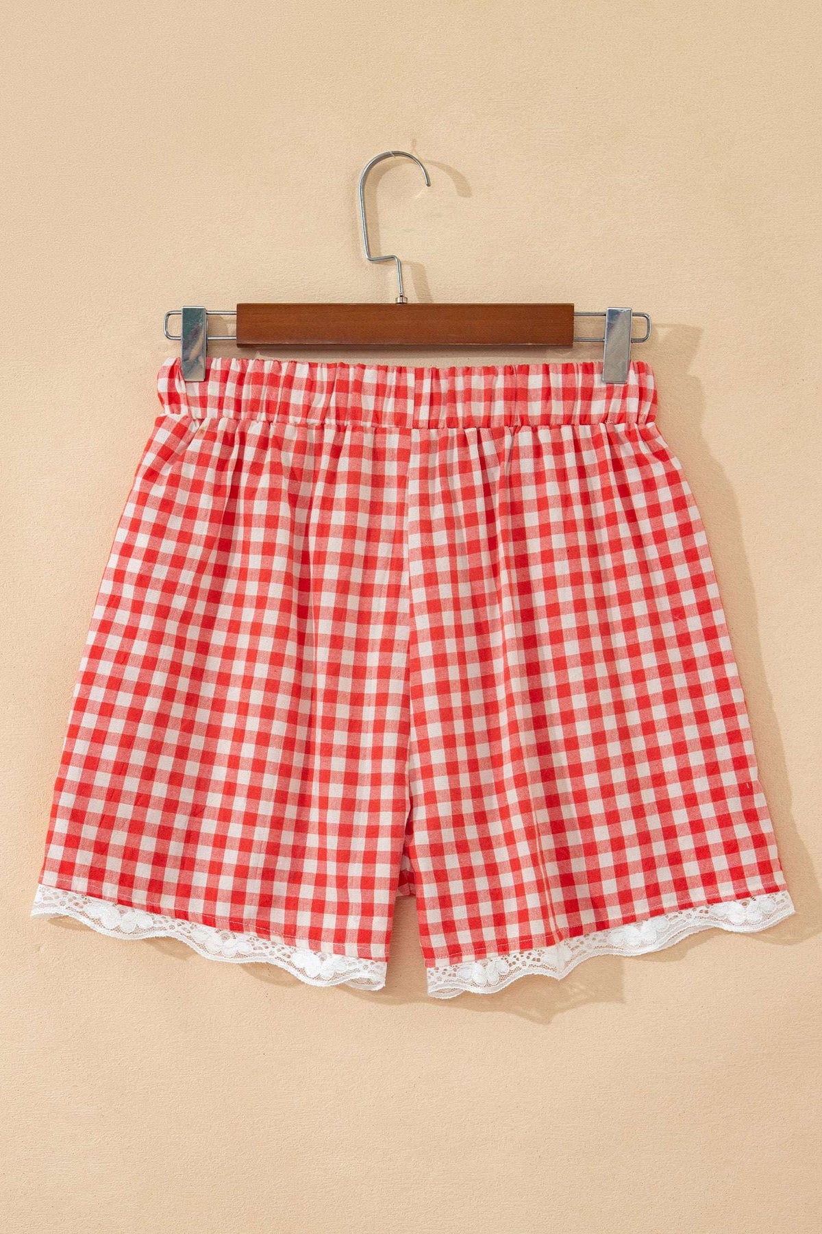 Red and white gingham fabric shorts with an elastic waist and white lace trim at the hem, hanging on a wooden clip hanger.