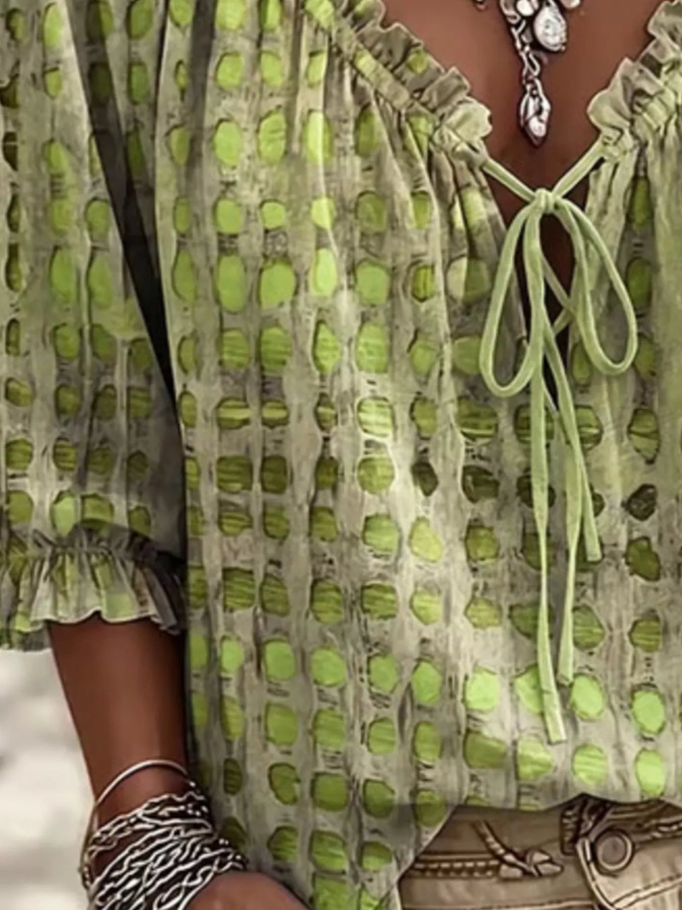 A close-up of a person wearing a light olive green and beige patterned blouse with a tie neckline, ruffled sleeves, and a textured dot design.