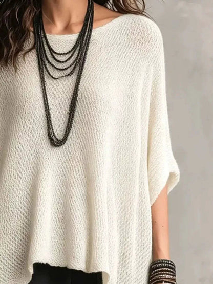 A close-up of a person wearing a loose, off-white textured knit top with short wide sleeves, layered with multiple long dark necklaces and bracelets, against a neutral gray background.