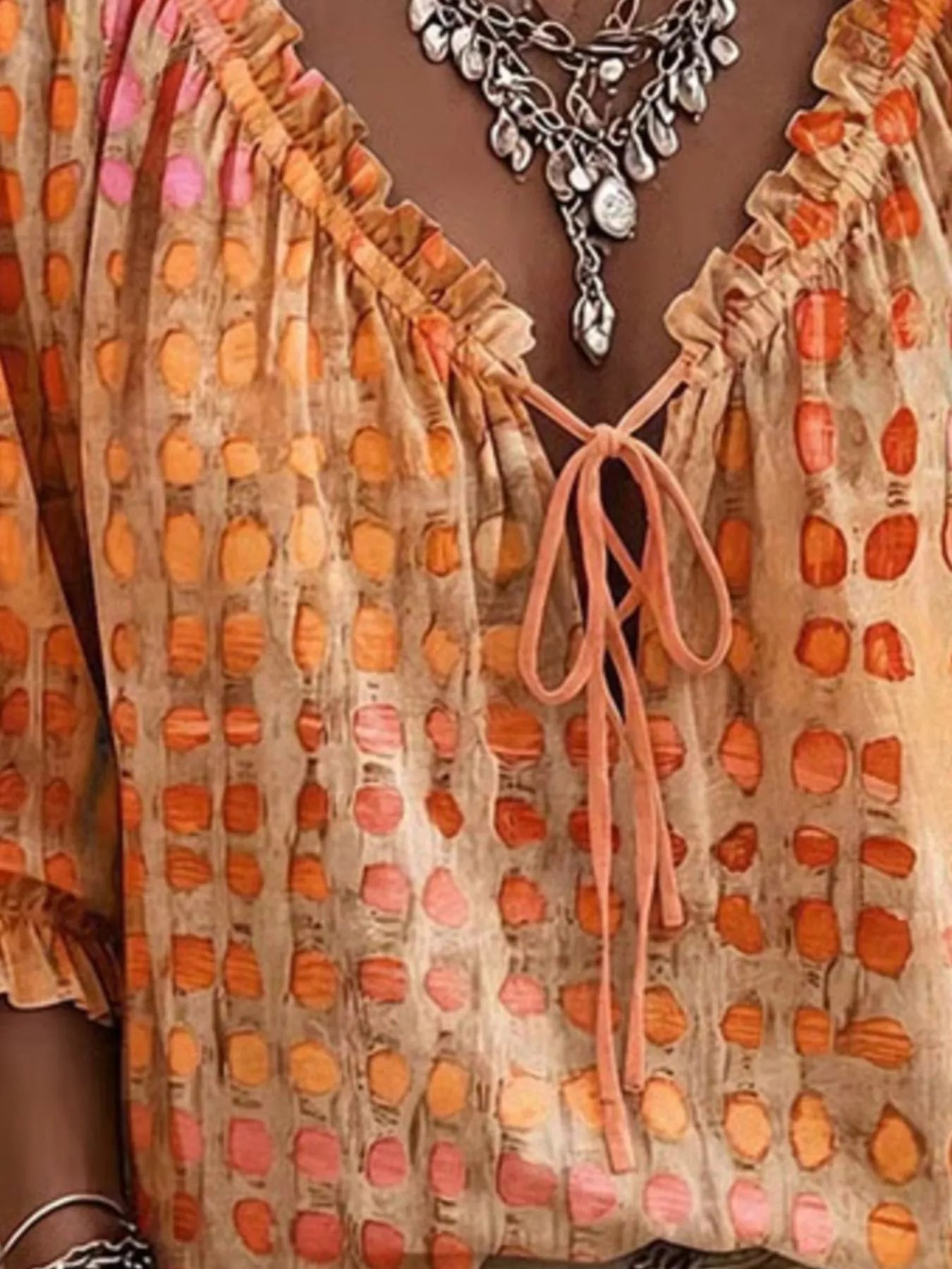 Close-up of a woman wearing a beige blouse with orange and pink abstract oval patterns, ruffled neckline, and tie-front detail, paired with layered silver necklaces.