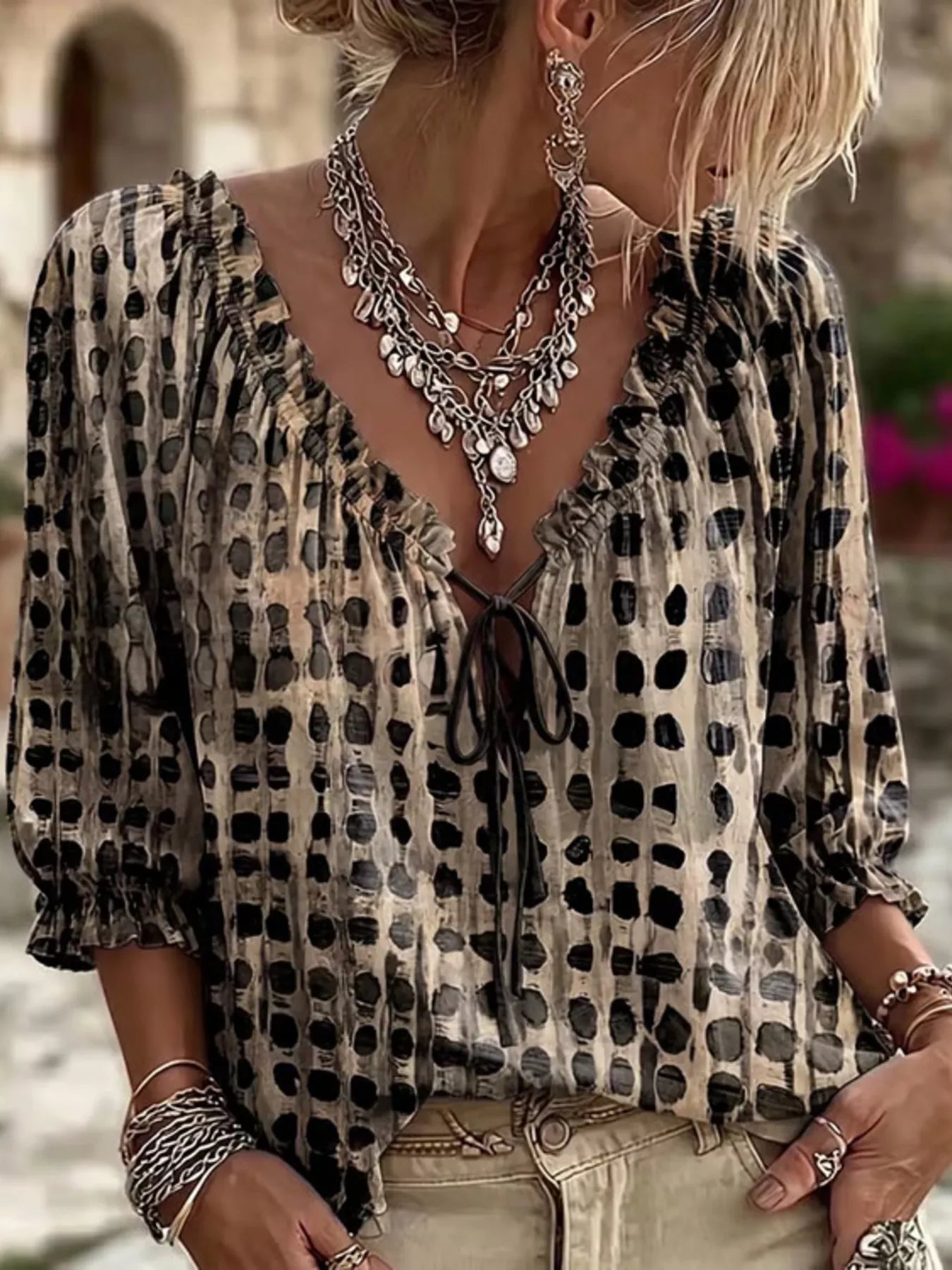 A woman wearing a beige and black leopard-print blouse with a ruffled V-neckline and tie-front detail, styled with layered silver statement necklaces and bracelets.