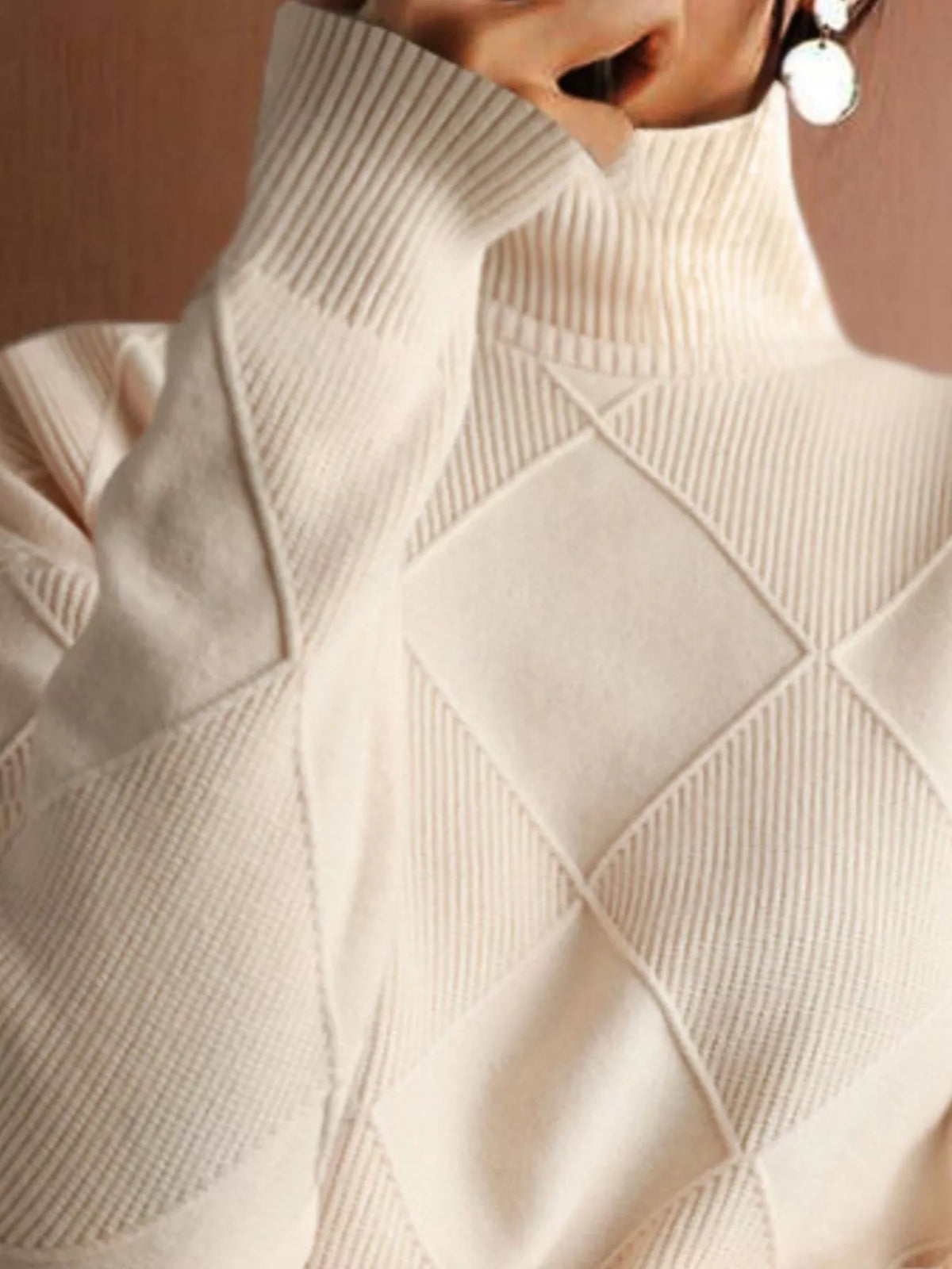 Close-up of a cream-colored knit sweater with a diamond argyle pattern and ribbed turtleneck, worn by a model.