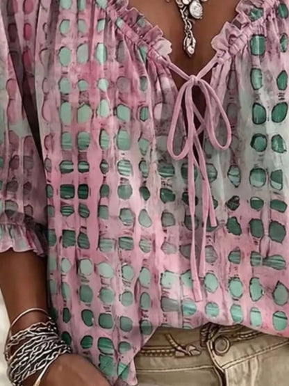 A close-up of a person wearing a pink and mint green patterned blouse with a ruffled V-neckline and tie-front detail, paired with beige pants and silver jewelry.