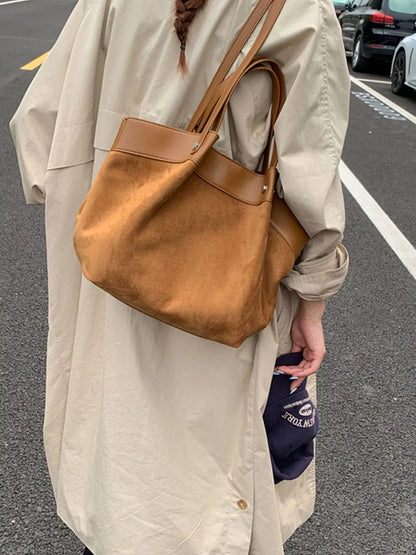 A person wearing a long beige coat carries a large brown suede tote bag on their shoulder in an outdoor street setting.