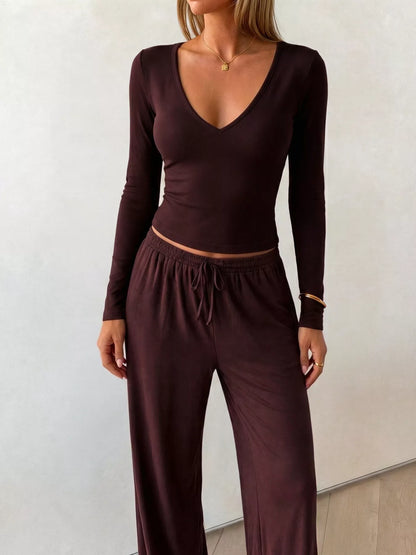 A woman wearing a matching dark plum-brown long-sleeve top and drawstring pants set, standing against a plain light wall.