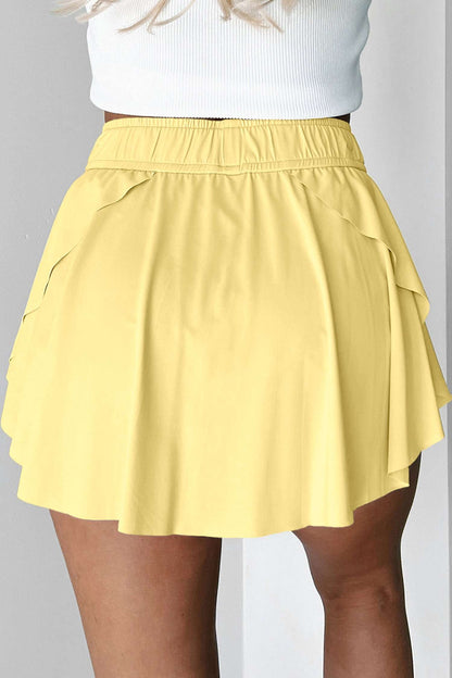 Yellow fabric tennis skort with an elastic waistband and ruffled hem shown from the back.