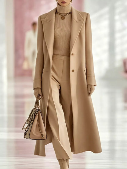 Model wearing a tan longline coat with a lapel collar and button front over a tan turtleneck and tan trousers.
