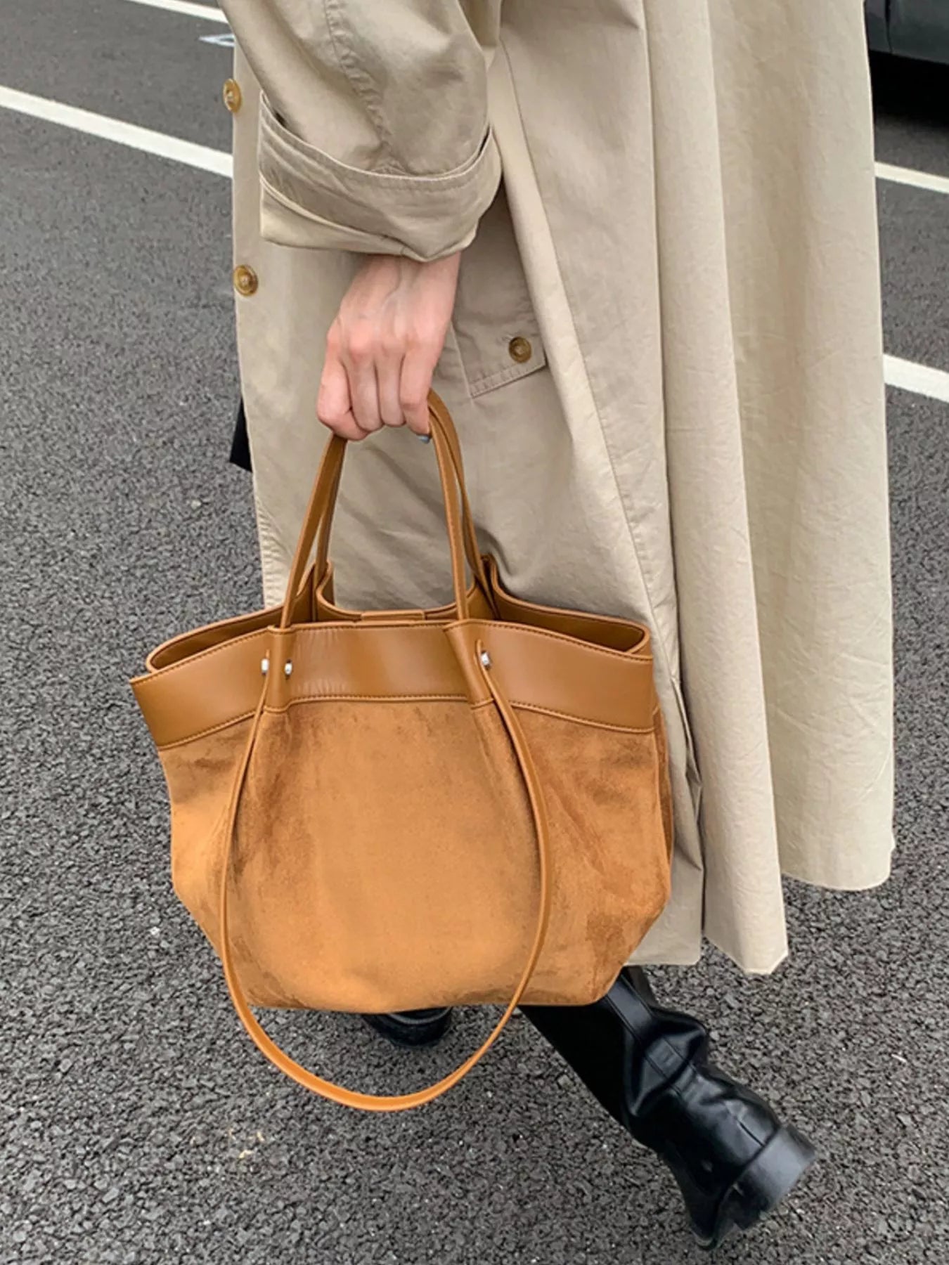 A person wearing a beige trench coat and black boots carries a large tan leather tote bag with double top handles and a shoulder strap while standing on asphalt.