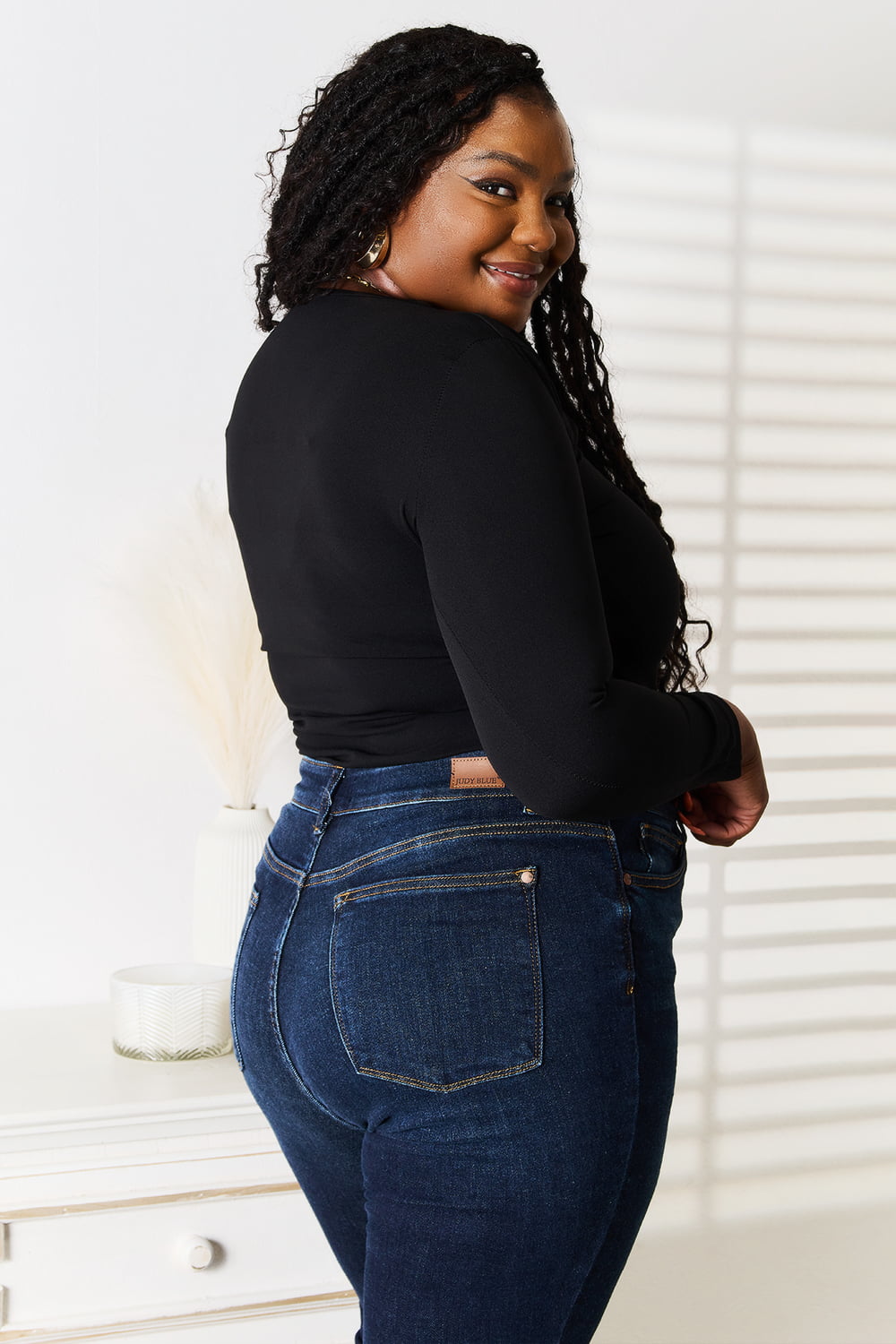 Woman wearing a black long-sleeve top and blue jeans standing in a room with white walls and furniture.
