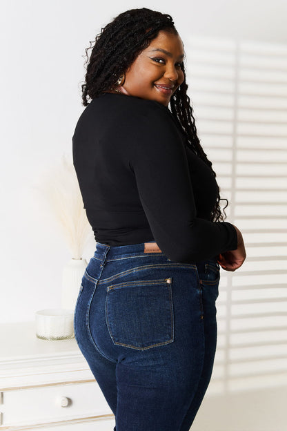 Woman wearing a black long-sleeve top and blue jeans standing in a room with white walls and furniture.