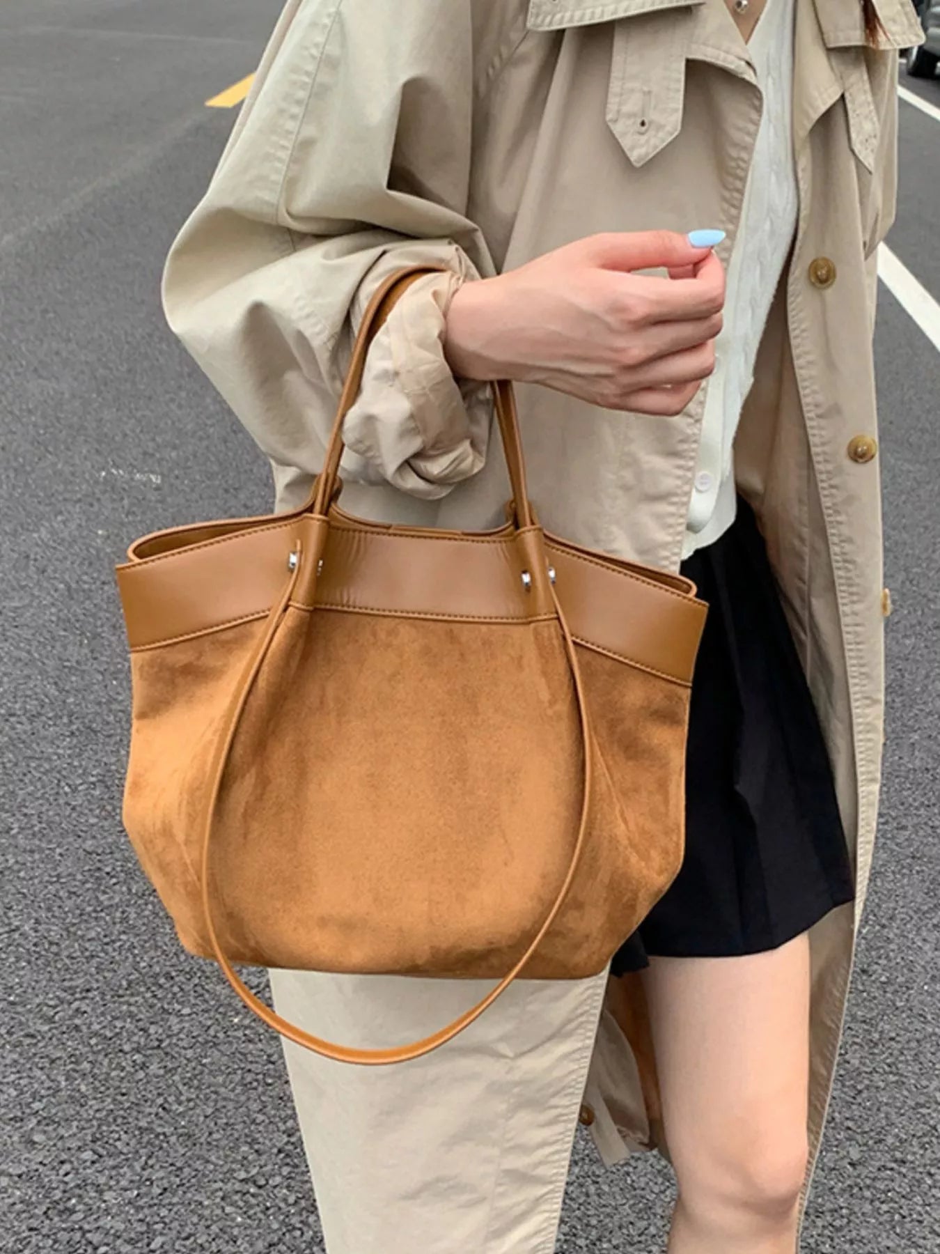 A person wearing a beige trench coat carries a large brown suede tote bag with matching leather trim and shoulder straps, photographed outdoors on a paved street.