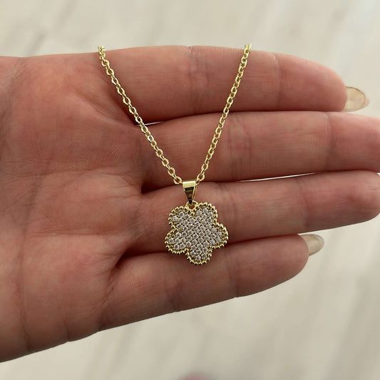 Gold necklace with a clover-shaped pendant held in a hand against a neutral background