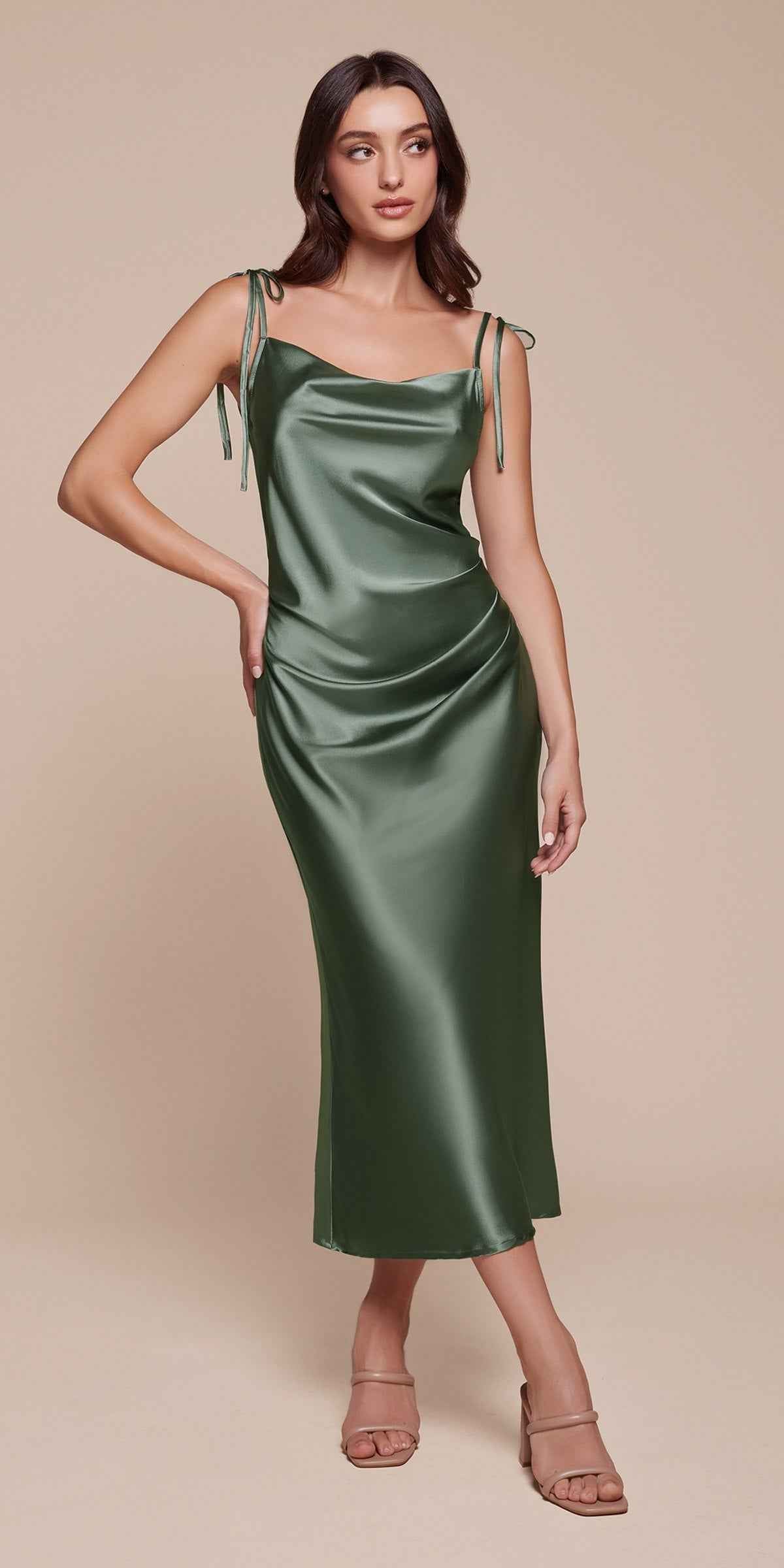 A woman modeling a satin slip dress in a muted sage green color, featuring thin shoulder straps with tie details and a draped, ruched bodice. She is wearing nude-colored strappy heels against a solid beige background.