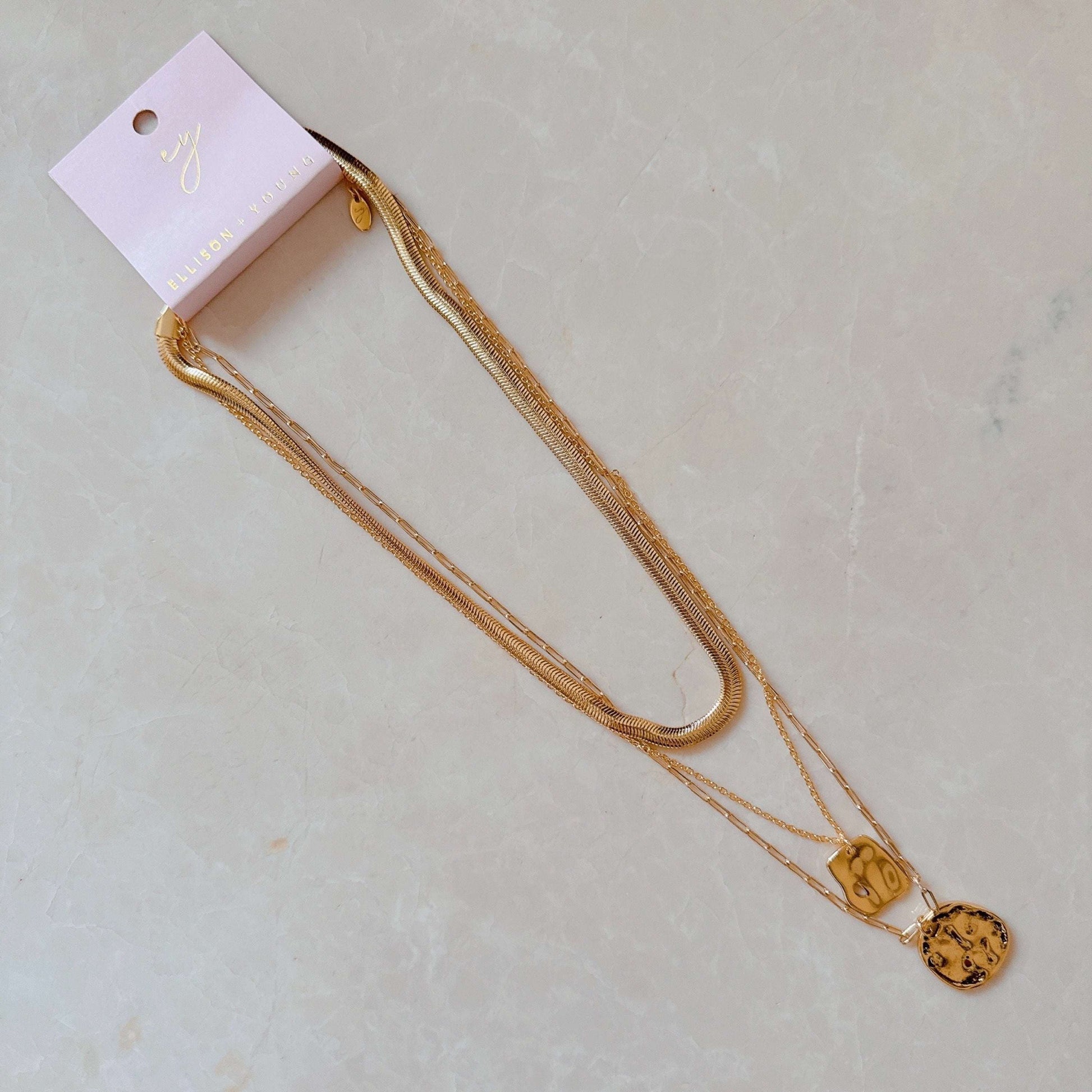 Three layered gold metal necklaces with two pendants on a pink ELLISON + YOUNG card.