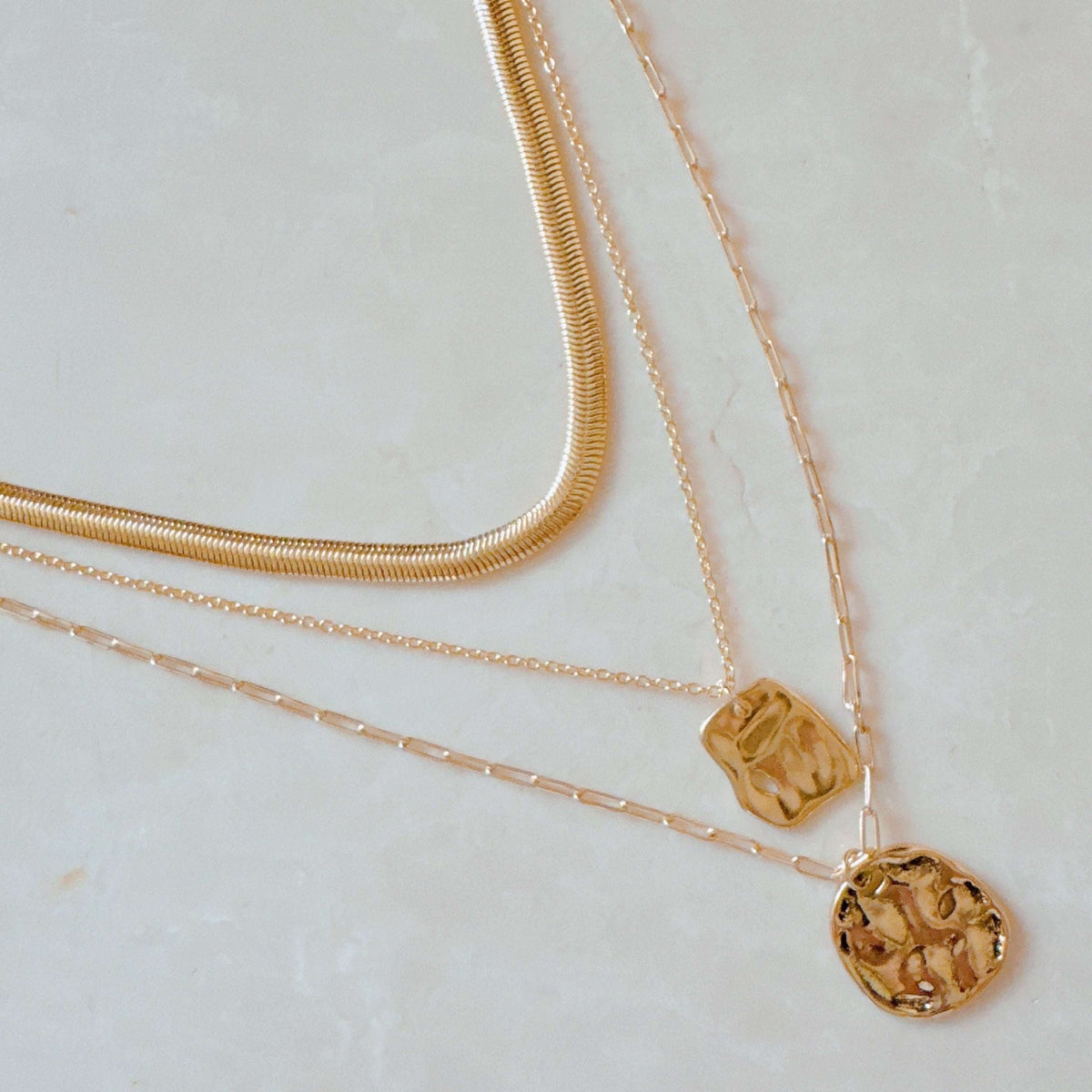 Three layered gold metal necklaces including a flat snake chain and two pendant chains with hammered square and round charms.
