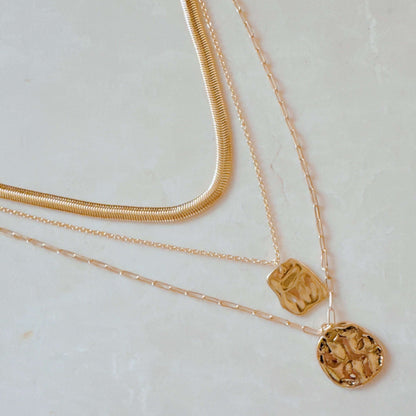 Three layered gold metal necklaces including a flat snake chain and two pendant chains with hammered square and round charms.