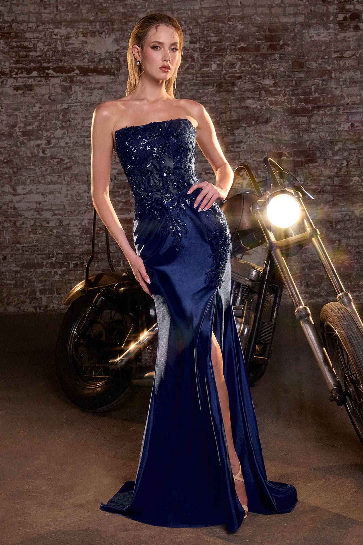 Woman in a strapless blue evening gown standing next to a motorcycle with a brick wall background