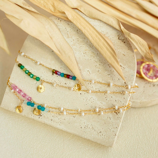 Set of gold bracelets with colorful beads on a textured surface with dried leaves.