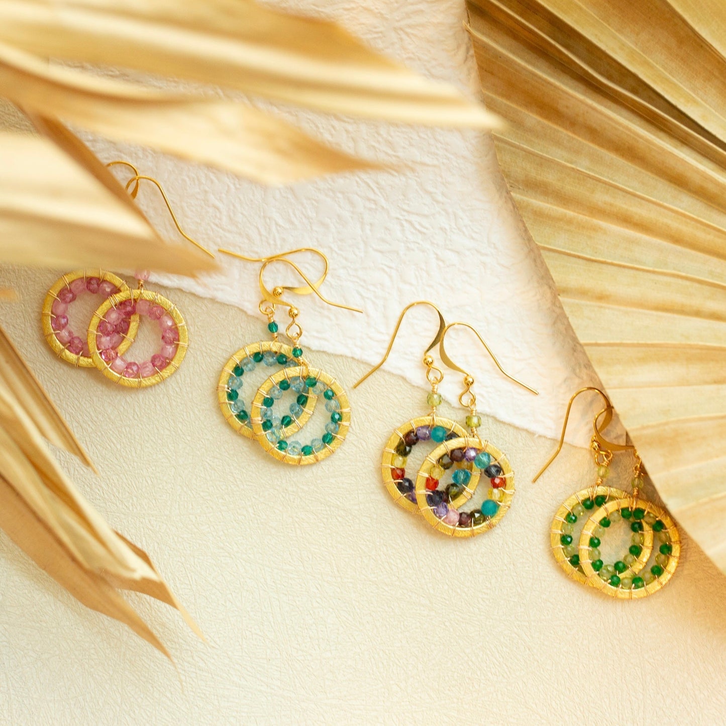 Gold earrings with colorful gemstones on a textured beige surface with gold leaves.
