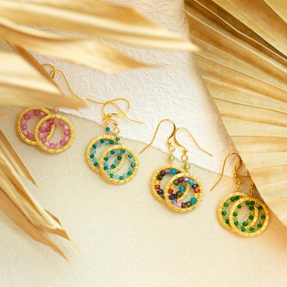 Gold earrings with colorful gemstones on a textured beige surface with gold leaves.