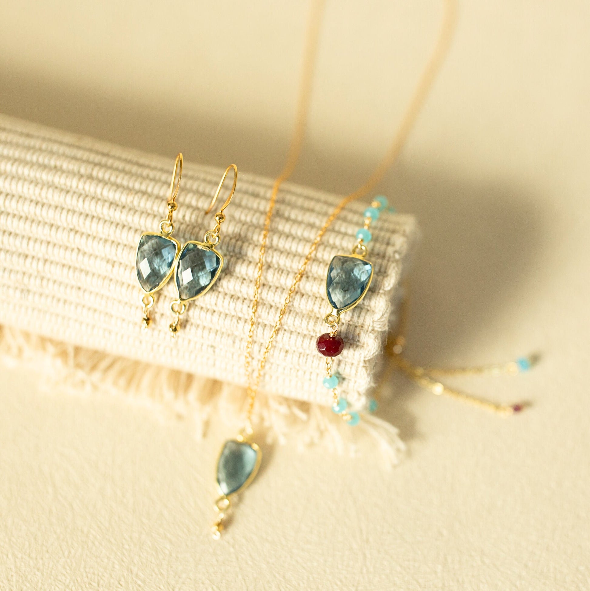 Gold jewelry set featuring a necklace and matching drop earrings with light blue marquise-cut gemstones, displayed on a cream textured fabric roll.