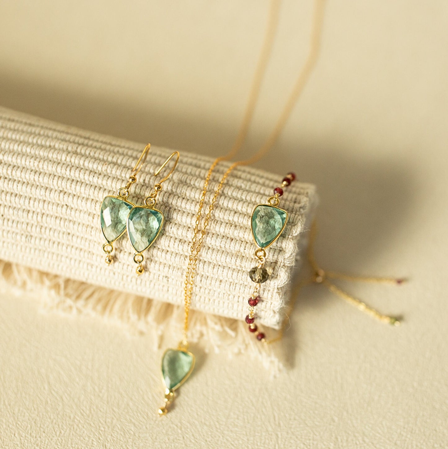 Gold jewelry set featuring heart-shaped green gemstone drop earrings, a matching pendant necklace, and a beaded bracelet, displayed on a textured cream fabric roll.