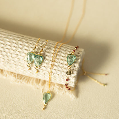 Gold jewelry set featuring heart-shaped green gemstone drop earrings, a matching pendant necklace, and a beaded bracelet, displayed on a textured cream fabric roll.