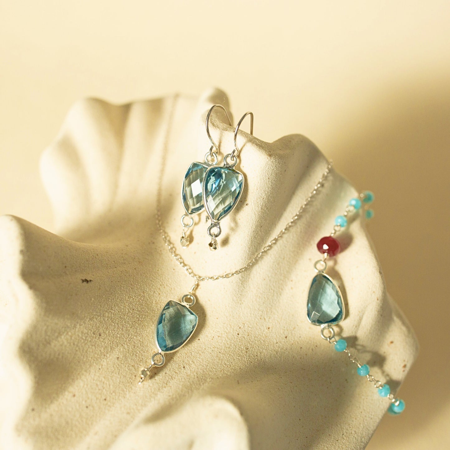 A jewelry set featuring a silver-toned necklace and matching drop earrings with faceted light blue teardrop-shaped gemstones, displayed on a textured cream-colored stand against a warm beige background.