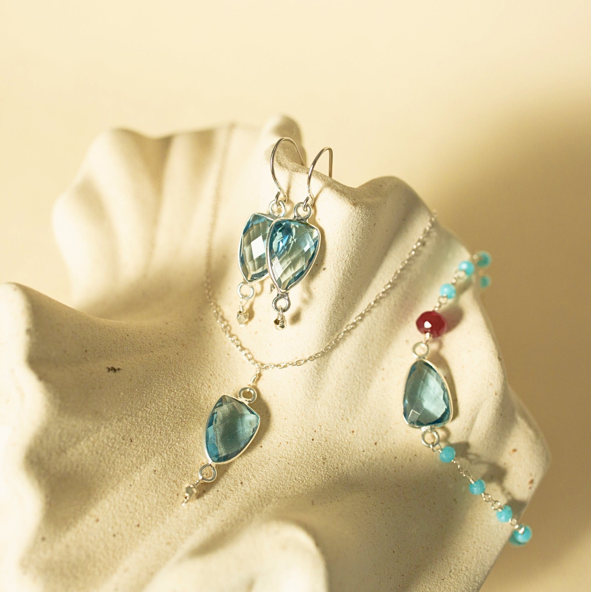 A jewelry set featuring a silver-toned necklace and matching drop earrings with faceted light blue teardrop-shaped gemstones, displayed on a textured cream-colored stand against a warm beige background.