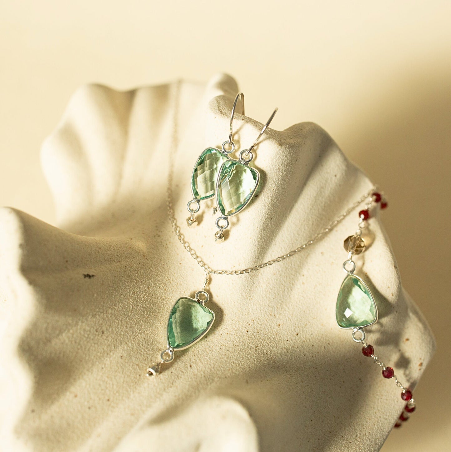 A jewelry set displayed on a cream-colored shell-shaped stand, featuring a silver necklace and matching earrings with light green teardrop-shaped gemstones and small red beads.