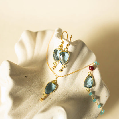 Gold jewelry set with light blue teardrop-shaped gemstones, including a necklace and matching earrings, displayed on a textured white shell against a warm beige background.