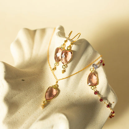 Gold-toned jewelry set displayed on a cream ceramic stand, featuring a necklace, matching earrings, and a bracelet with faceted blush pink stones and small red and blue accent beads.