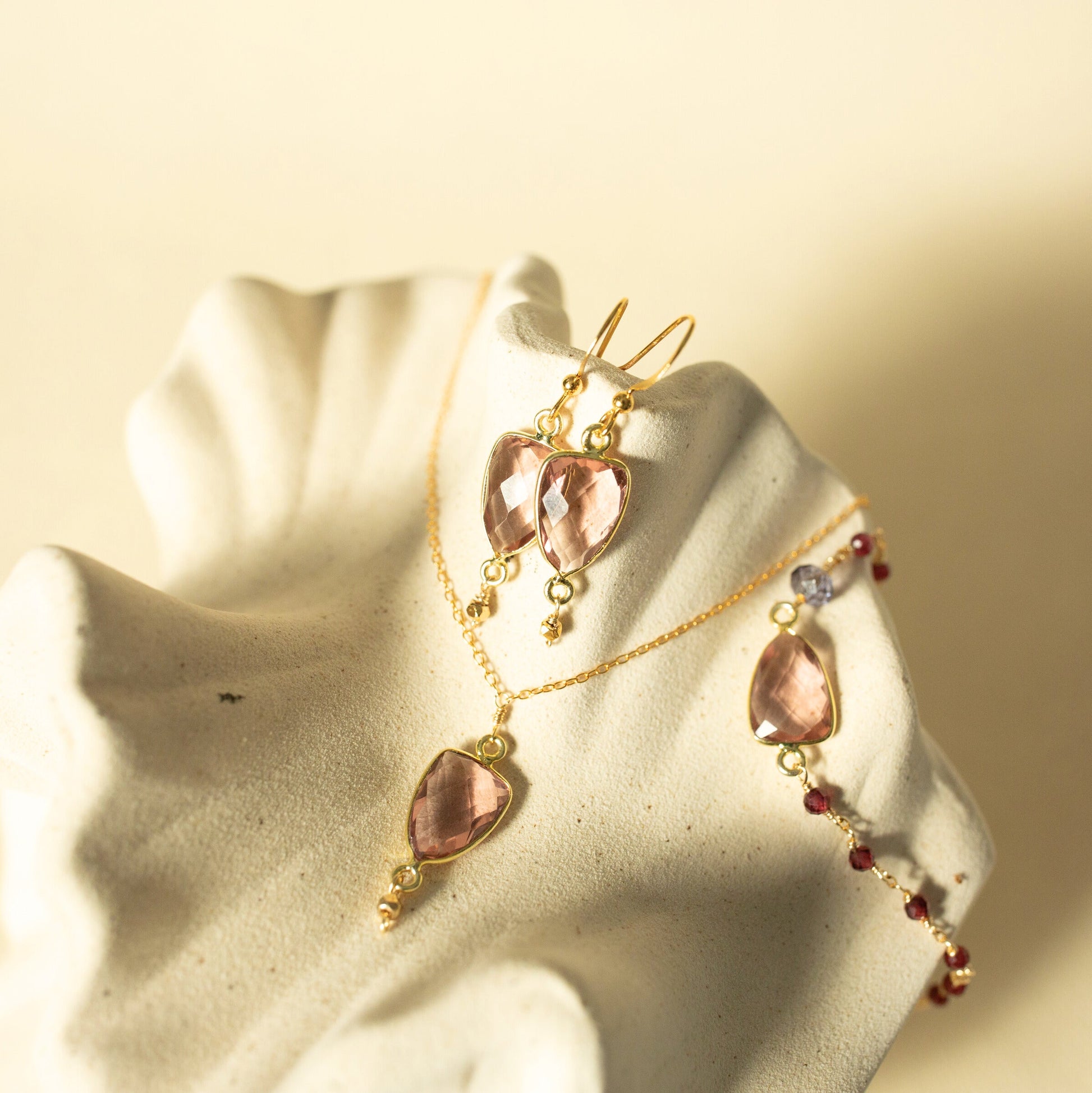Gold-toned jewelry set with large blush pink teardrop gemstones, including a necklace, earrings, and a bracelet, displayed on a textured cream-colored stand against a warm beige background.