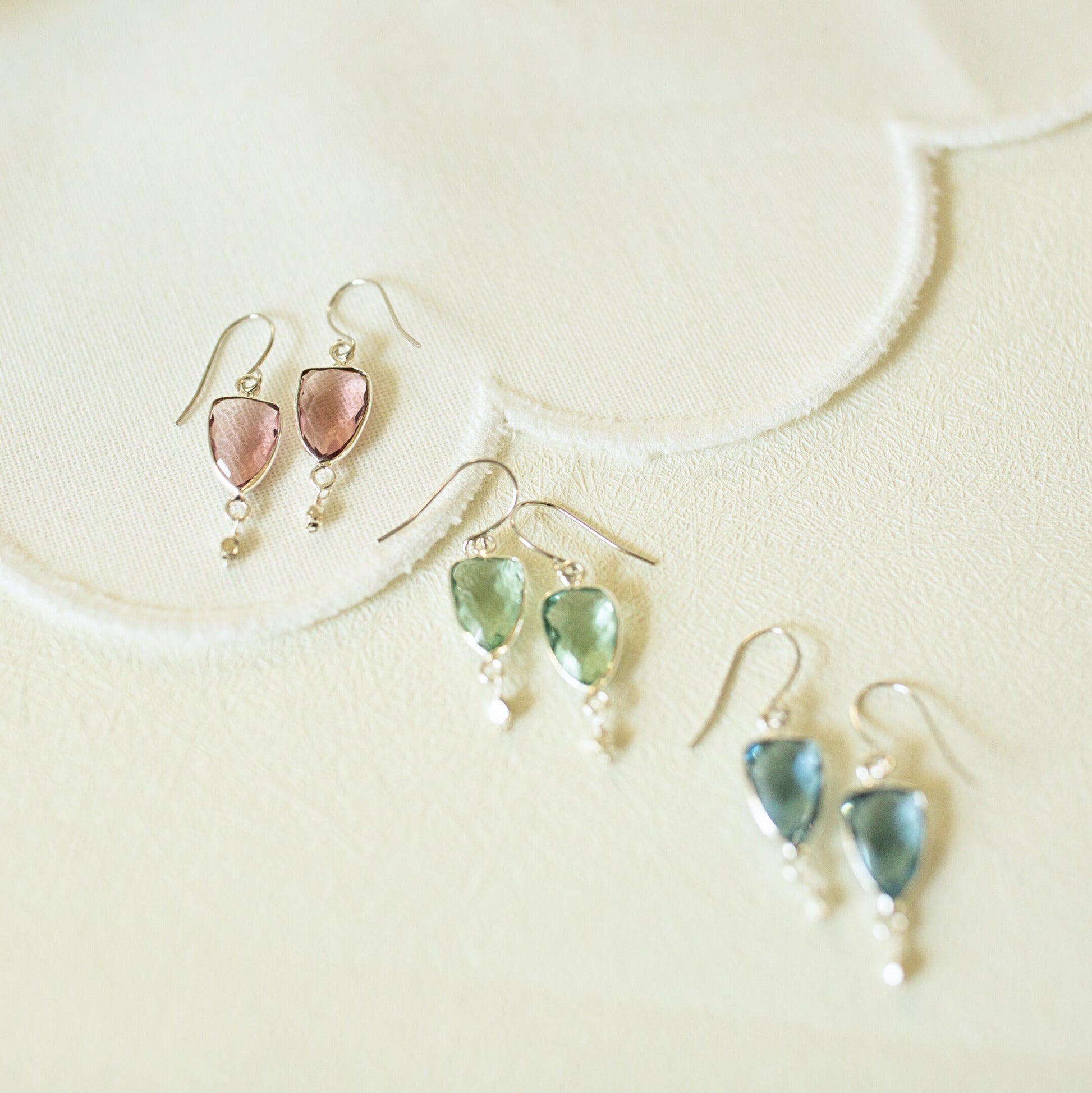 Three pairs of dangling earrings arranged on a light cream textured surface. Each pair has a small silver-tone fishhook and a teardrop-shaped gemstone drop with a tiny bead below.