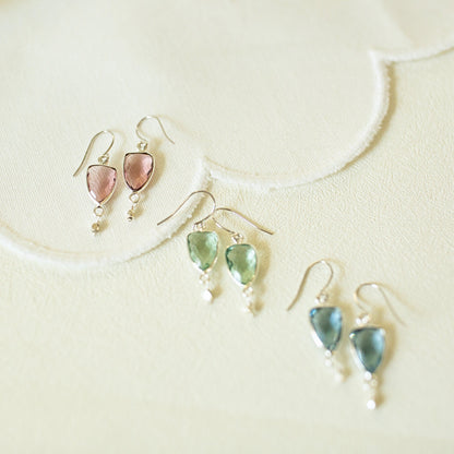 Three pairs of dangling earrings arranged on a light cream textured surface. Each pair has a small silver-tone fishhook and a teardrop-shaped gemstone drop with a tiny bead below.