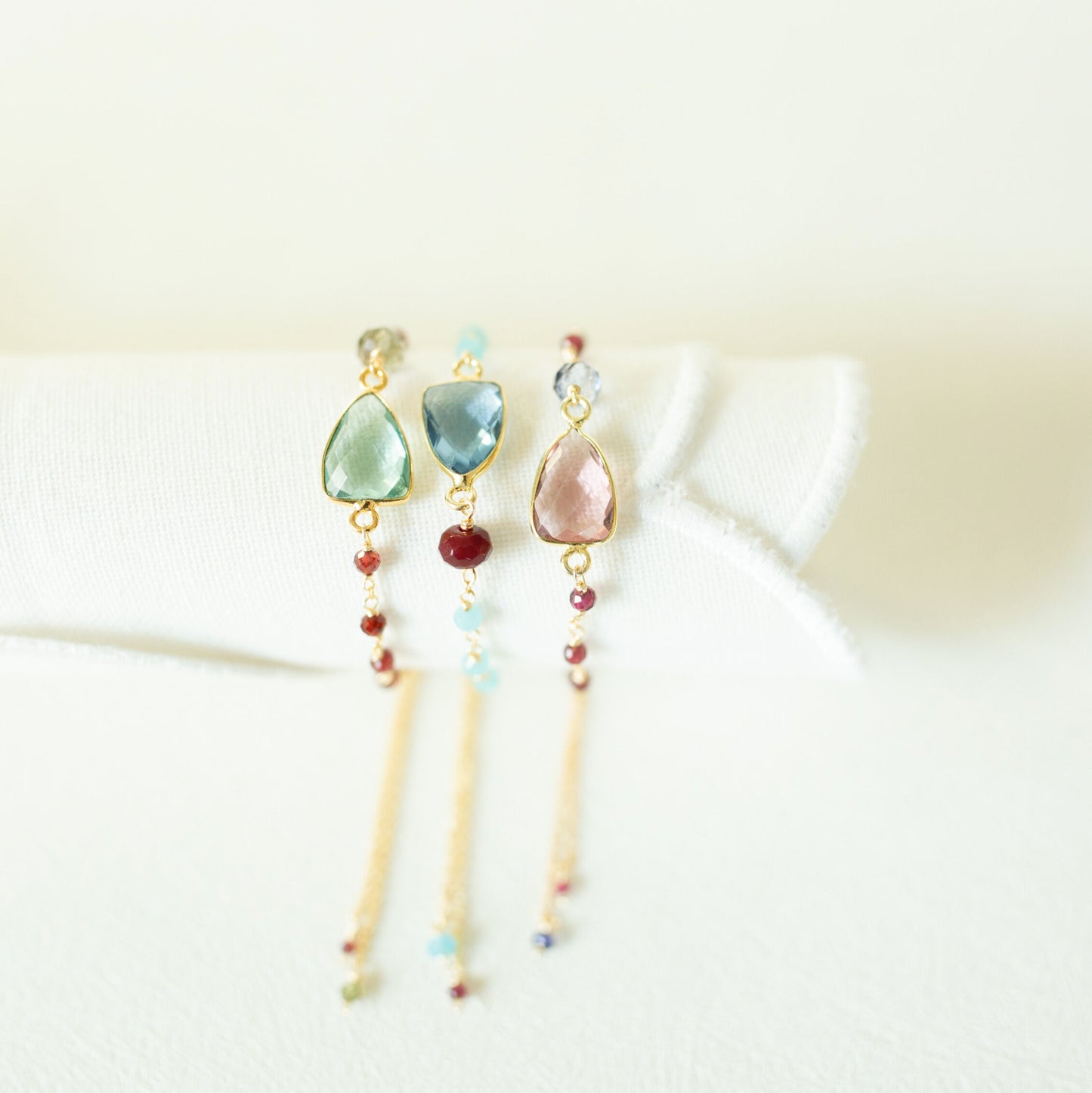 Three delicate gold-tone dangle earrings with teardrop-shaped gemstones in light green, light blue, and pale pink, accented with small round beads, displayed on a soft white fabric stand.