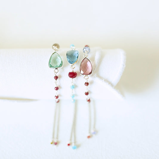 Three delicate dangle earrings displayed on a white fabric stand against a soft white background. Each earring features a teardrop-shaped gemstone in green, blue, and pink, with small red beads and silver-toned dangling chains.