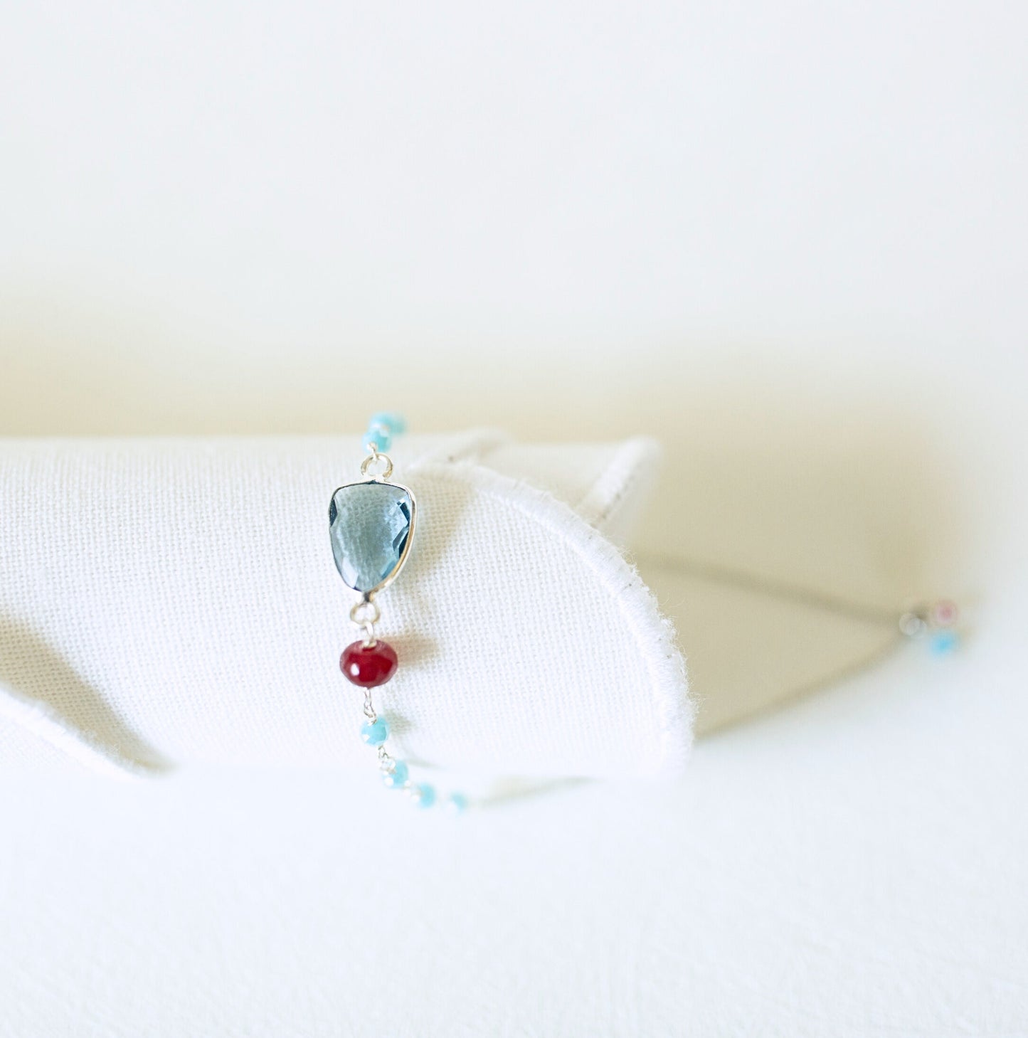 A delicate silver-toned bracelet displayed on a white fabric stand, featuring a faceted light blue gemstone, a single red bead, and small light blue beads.