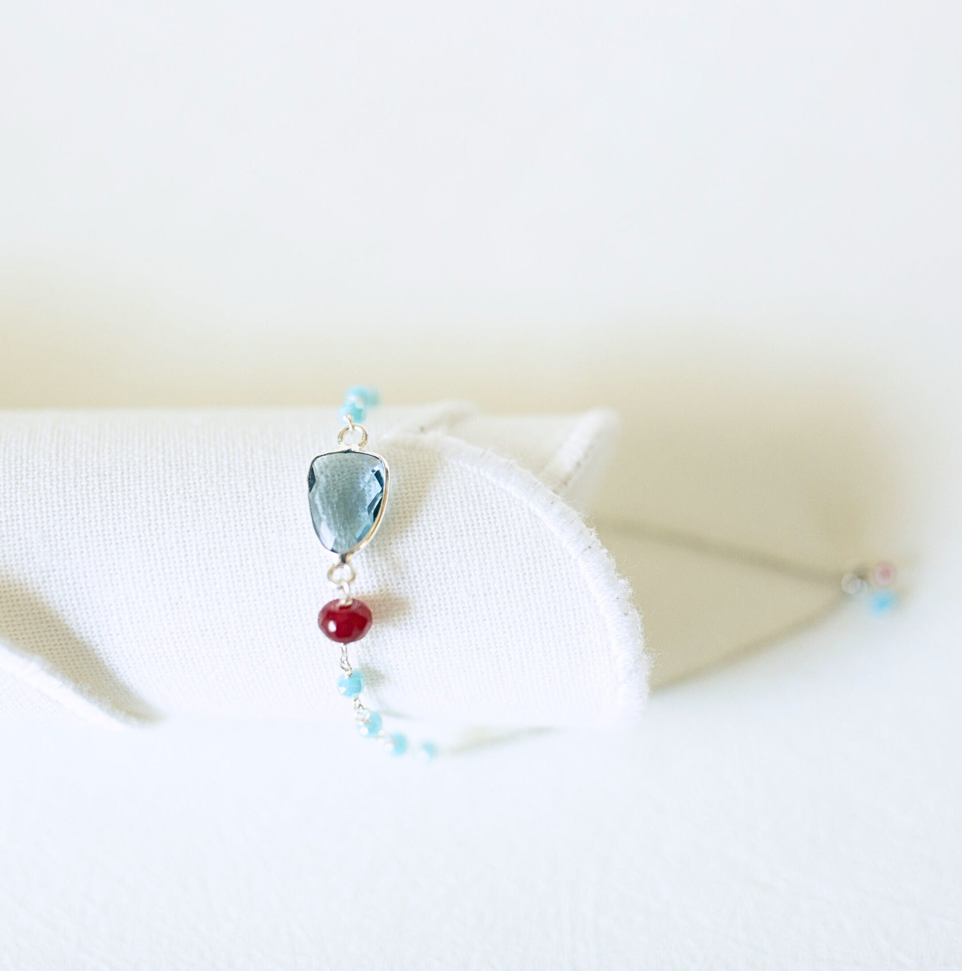 A delicate silver-toned bracelet displayed on a white fabric stand, featuring a faceted light blue gemstone, a single red bead, and small light blue beads.