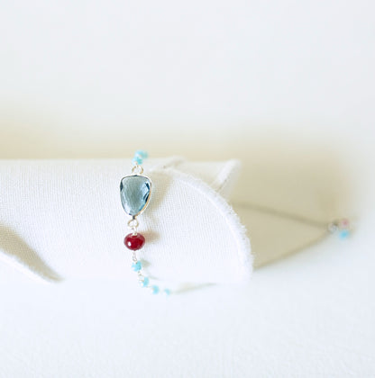 A delicate silver-toned bracelet displayed on a white fabric stand, featuring a faceted light blue gemstone, a single red bead, and small light blue beads.