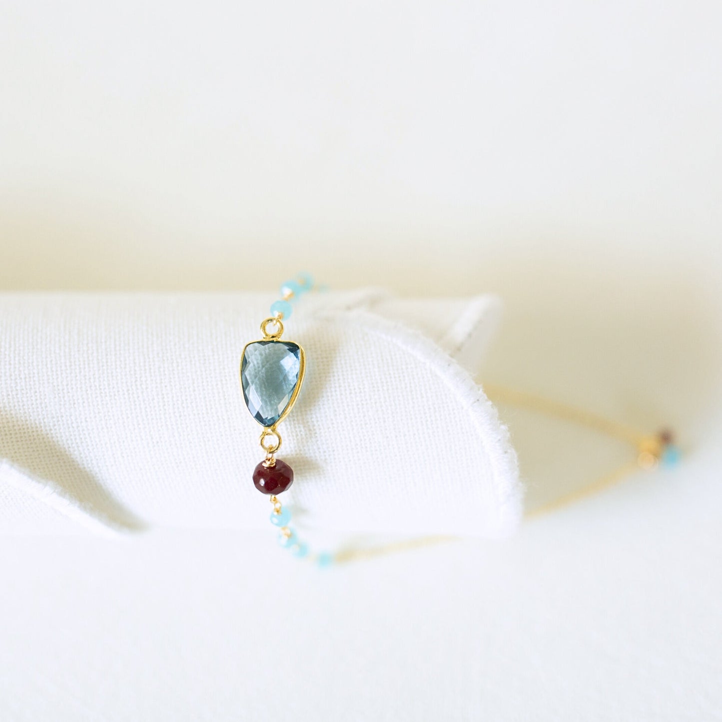 A delicate gold bracelet displayed on a white fabric stand, featuring a light blue marquise-cut gemstone, a small dark red bead, and tiny light blue beads.