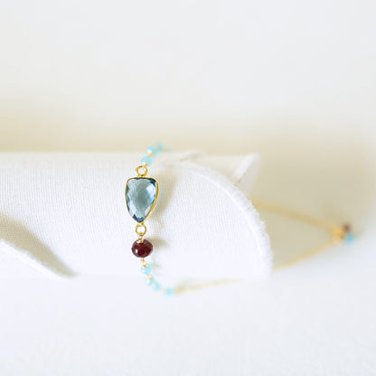 A delicate gold bracelet displayed on a white fabric stand, featuring a light blue marquise-cut gemstone, a small dark red bead, and tiny light blue beads.