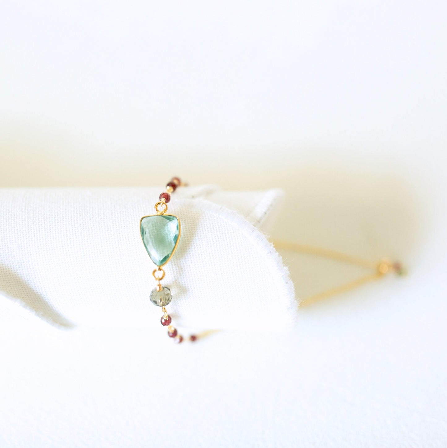 A delicate gold-tone beaded bracelet with a light green teardrop-shaped gemstone and a small gray bead, displayed on a white fabric stand.