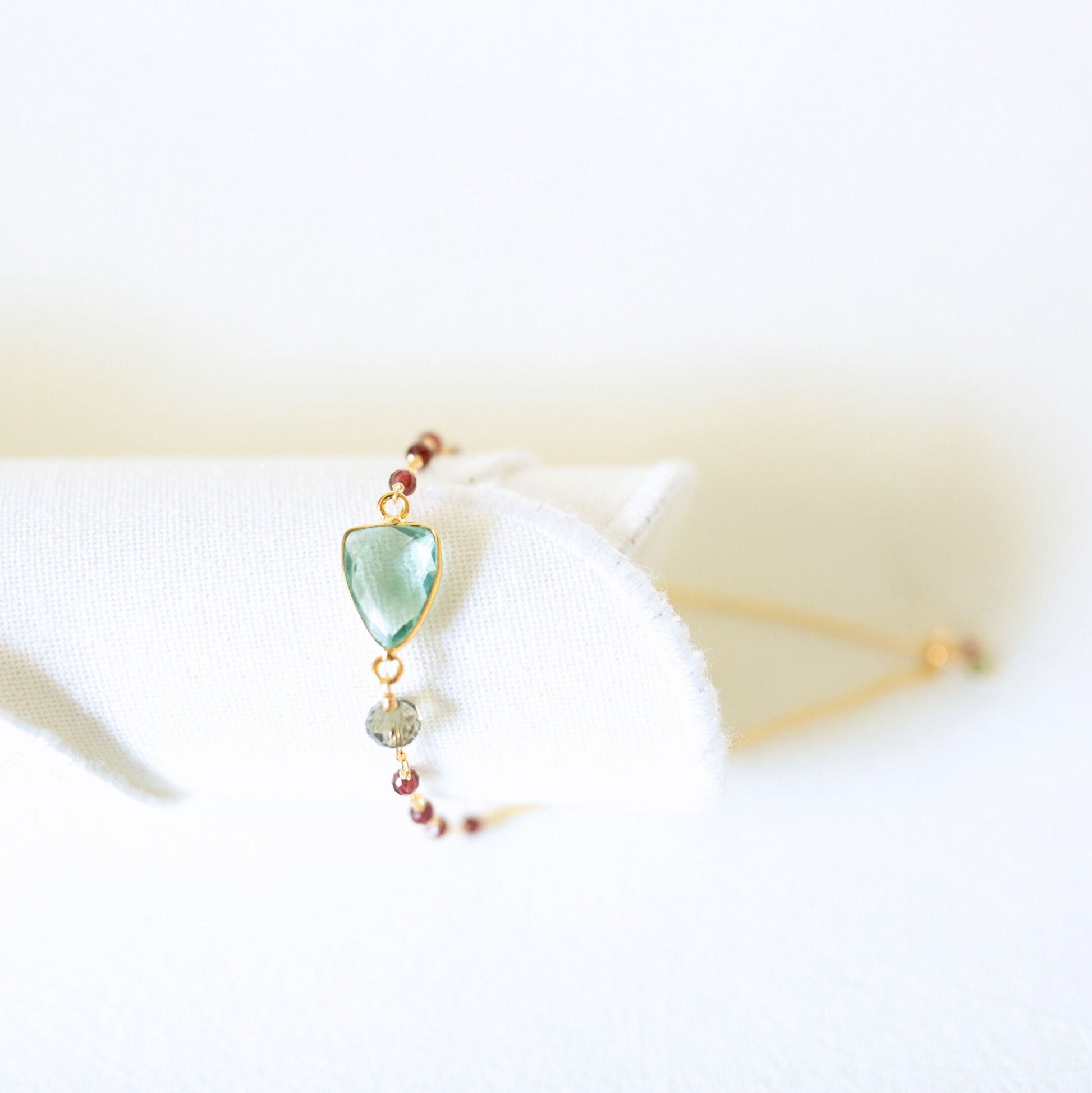 A delicate gold-tone beaded bracelet with a light green teardrop-shaped gemstone and a small gray bead, displayed on a white fabric stand.