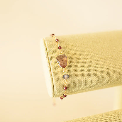 A delicate gold bracelet with a central brownish stone and small dark red beads, displayed on a textured beige fabric jewelry stand against a soft cream background.