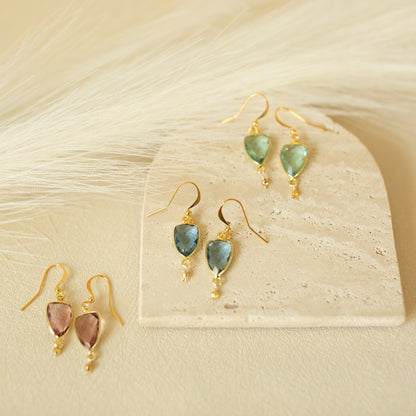 Three pairs of gold-tone dangle earrings with teardrop-shaped gemstones are arranged on a light stone slab and soft cream faux-fur background. Two pairs have pale blue-green stones, and one pair has blush pink stones.