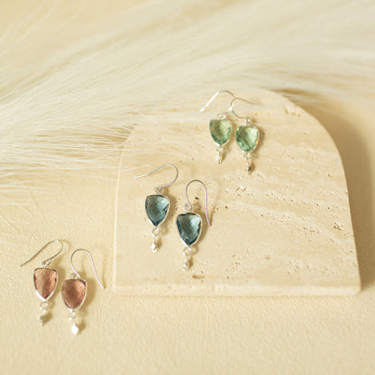 Four dangle earrings displayed on a light beige stone slab with soft cream-colored dried grass in the background. Two earrings feature dusty pink teardrop-shaped stones with silver hooks and bead drops, while the other two feature green teardrop-shaped stones with the same silver hardware.