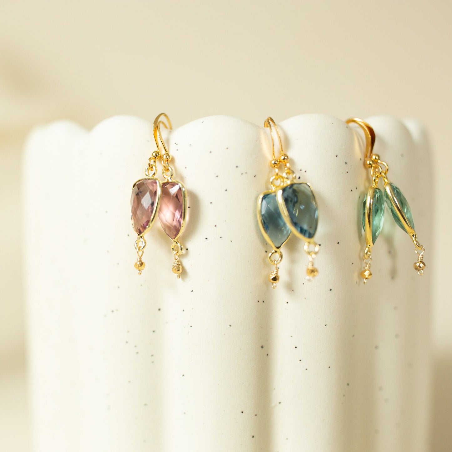Three pairs of gold dangle earrings with marquise-cut gemstones in pink, blue, and green, displayed on the scalloped edge of a white speckled ceramic pedestal.